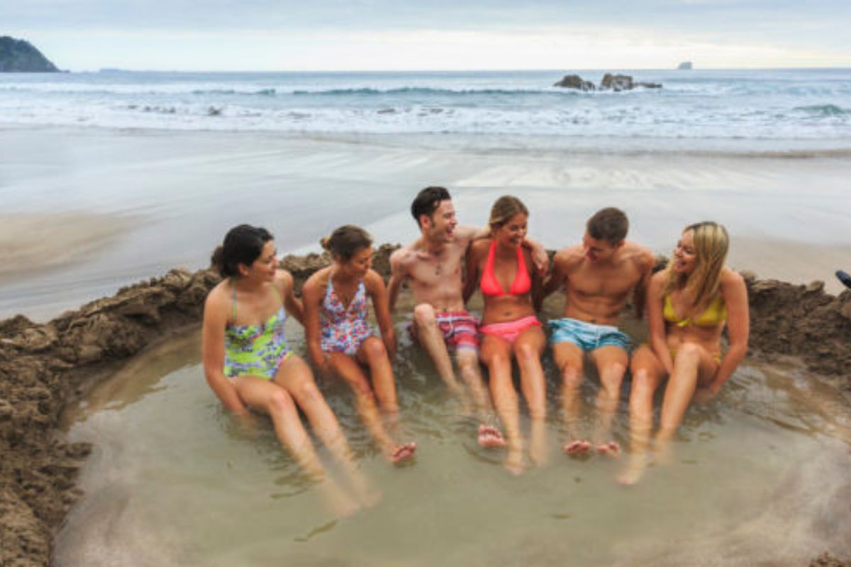 people in swimsuits sitting in a hot water beach pool at hot water beach in new zealand